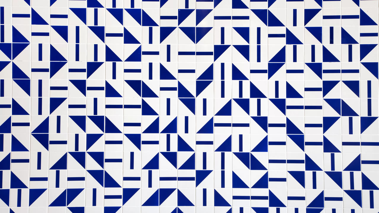 Parede de azulejos branco com grafismos azuis. Cada azulejo possui um desenho de triangulo ou retangulo e são colocados de forma aleatória gerando uma composição sem padrão aparente.