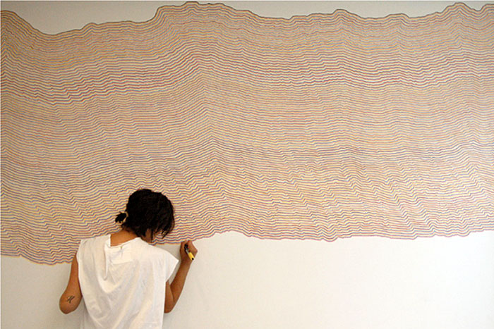 Uma pessoa de costa para a foto desenhando linhas coloridas consecutivas em uma parade branca. Ela está executando o WALL DRAWING 797 de Sol LeWitt.